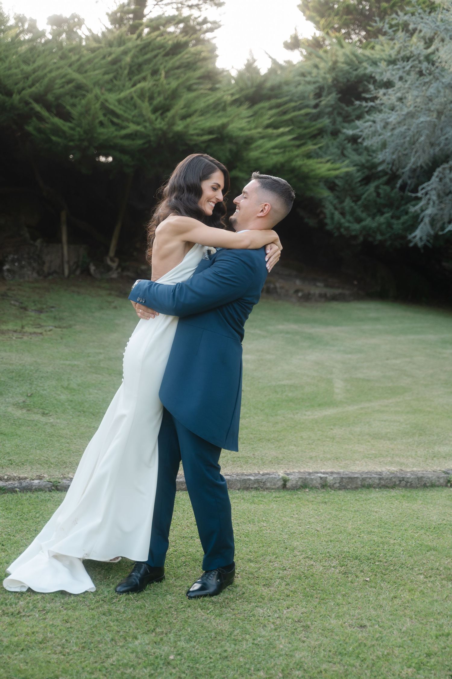A couple in wedding attire dances together on a grassy lawn with evergreen trees in the background.
