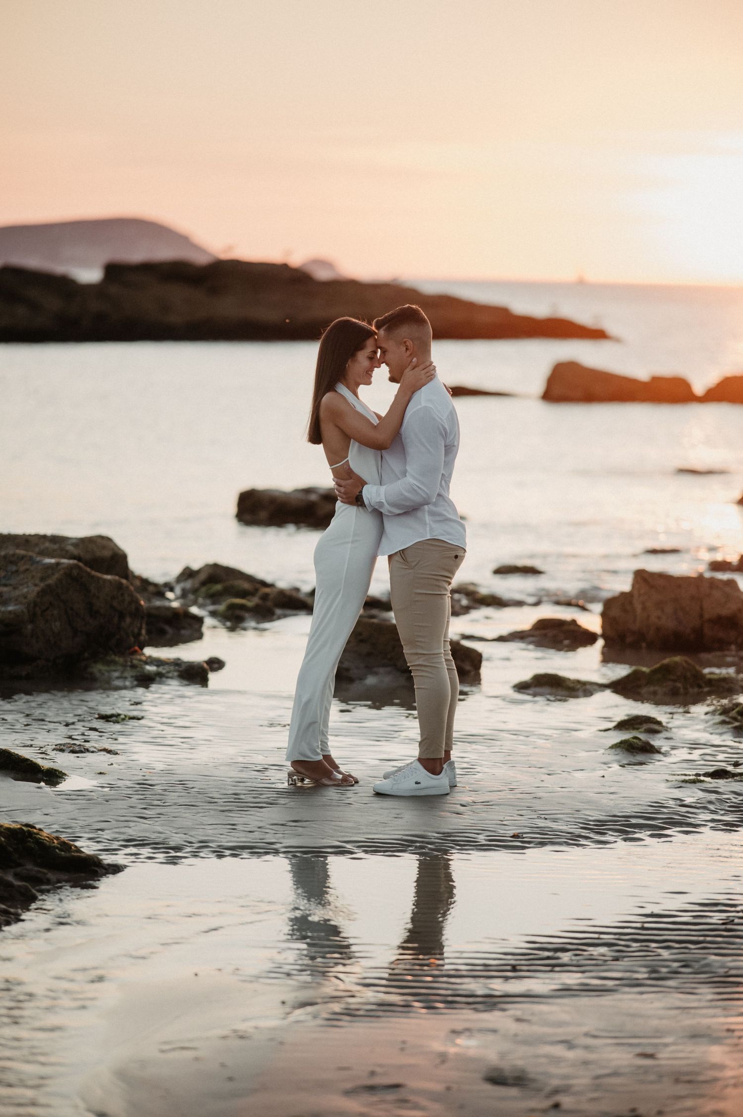 A couple stands together in shallow ocean water at sunset, surrounded by rocky coastline and golden light.