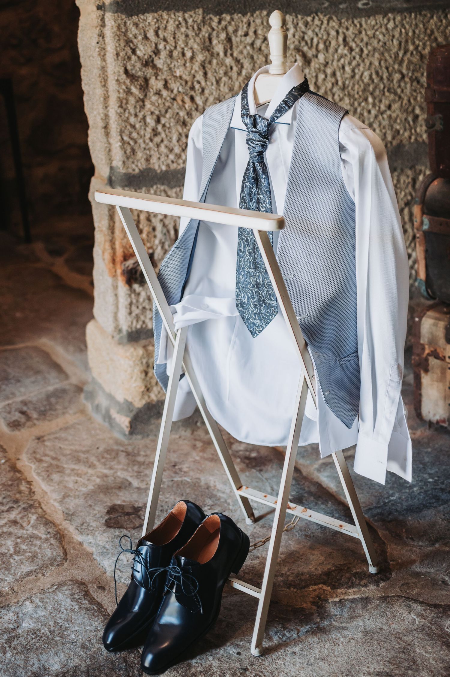 Formal white dress shirt, blue patterned tie, and black dress shoes arranged on a white clothing rack against a textured wall.
