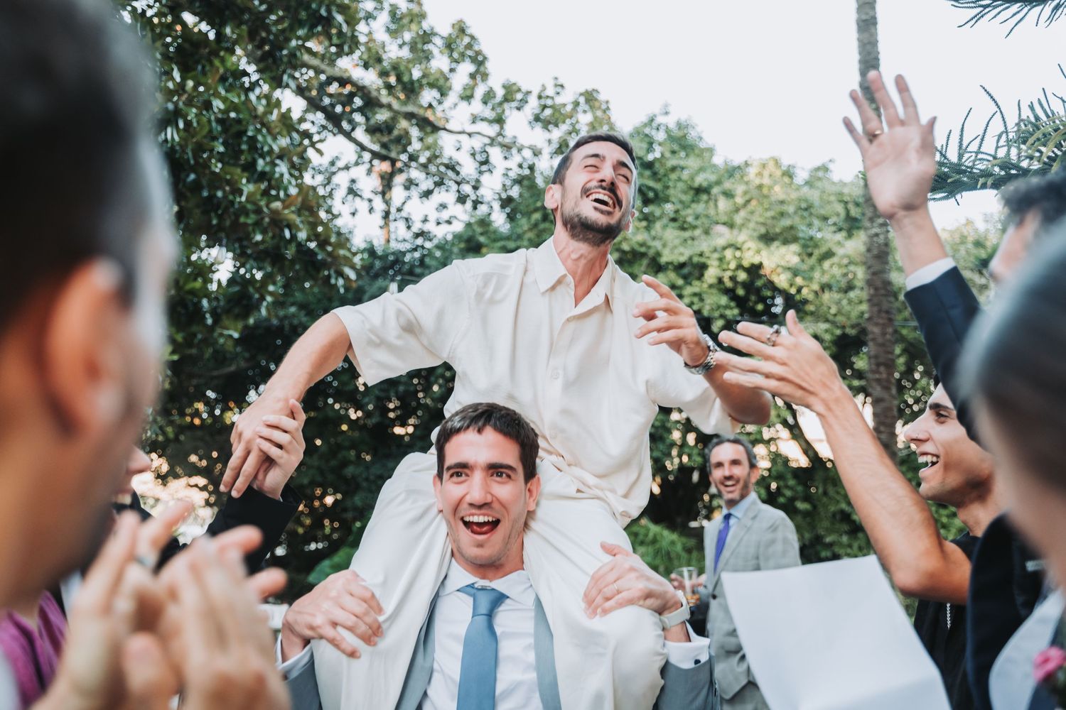 A joyful wedding celebration with guests dancing and raising their hands in an outdoor garden setting.