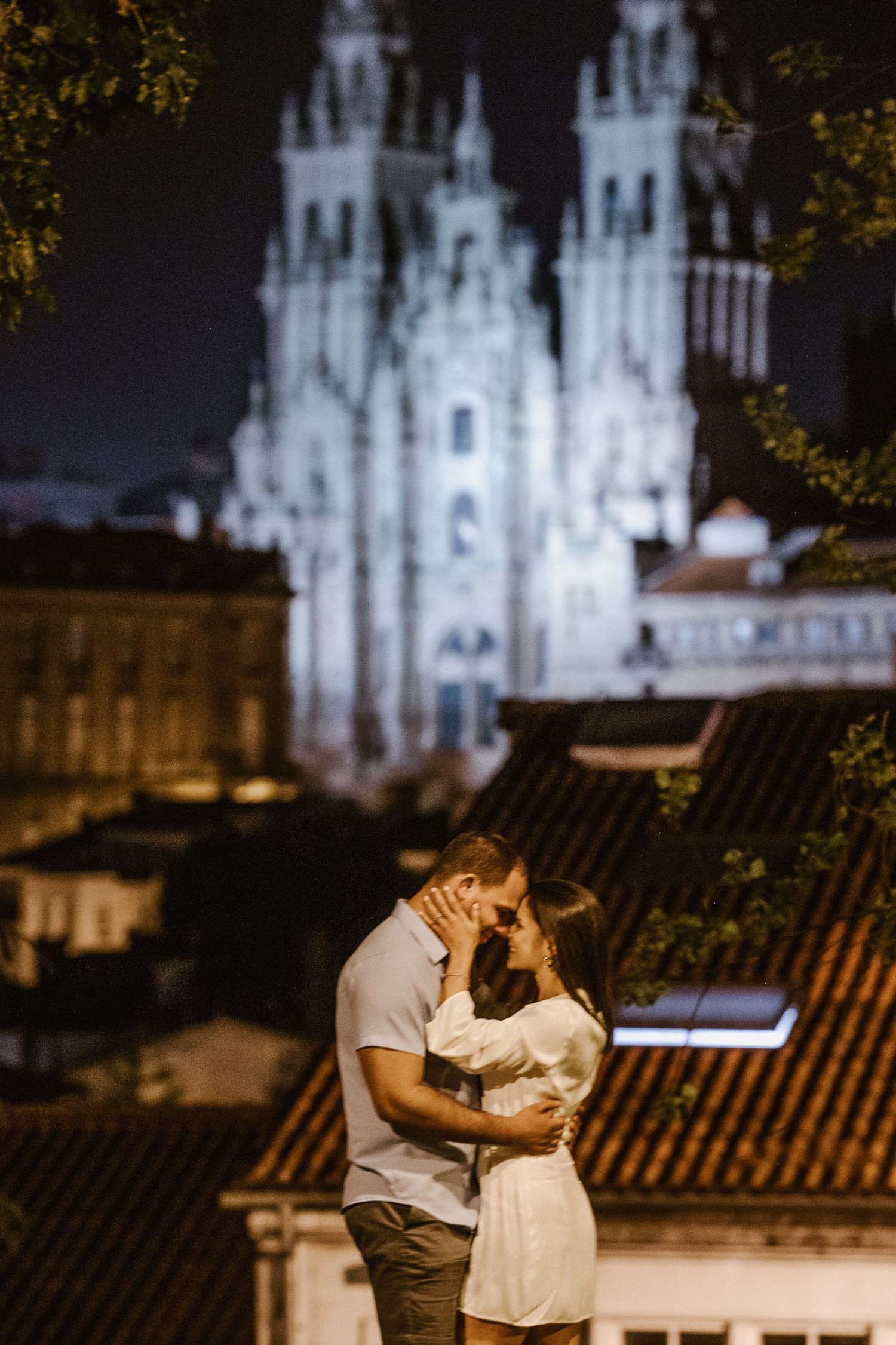 A romantic couple shares an intimate moment against a backdrop of illuminated cathedral spires at night.