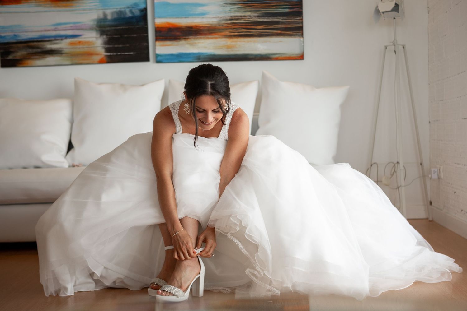 Bride in flowing white gown fastening her elegant wedding shoes in modern living room setting.