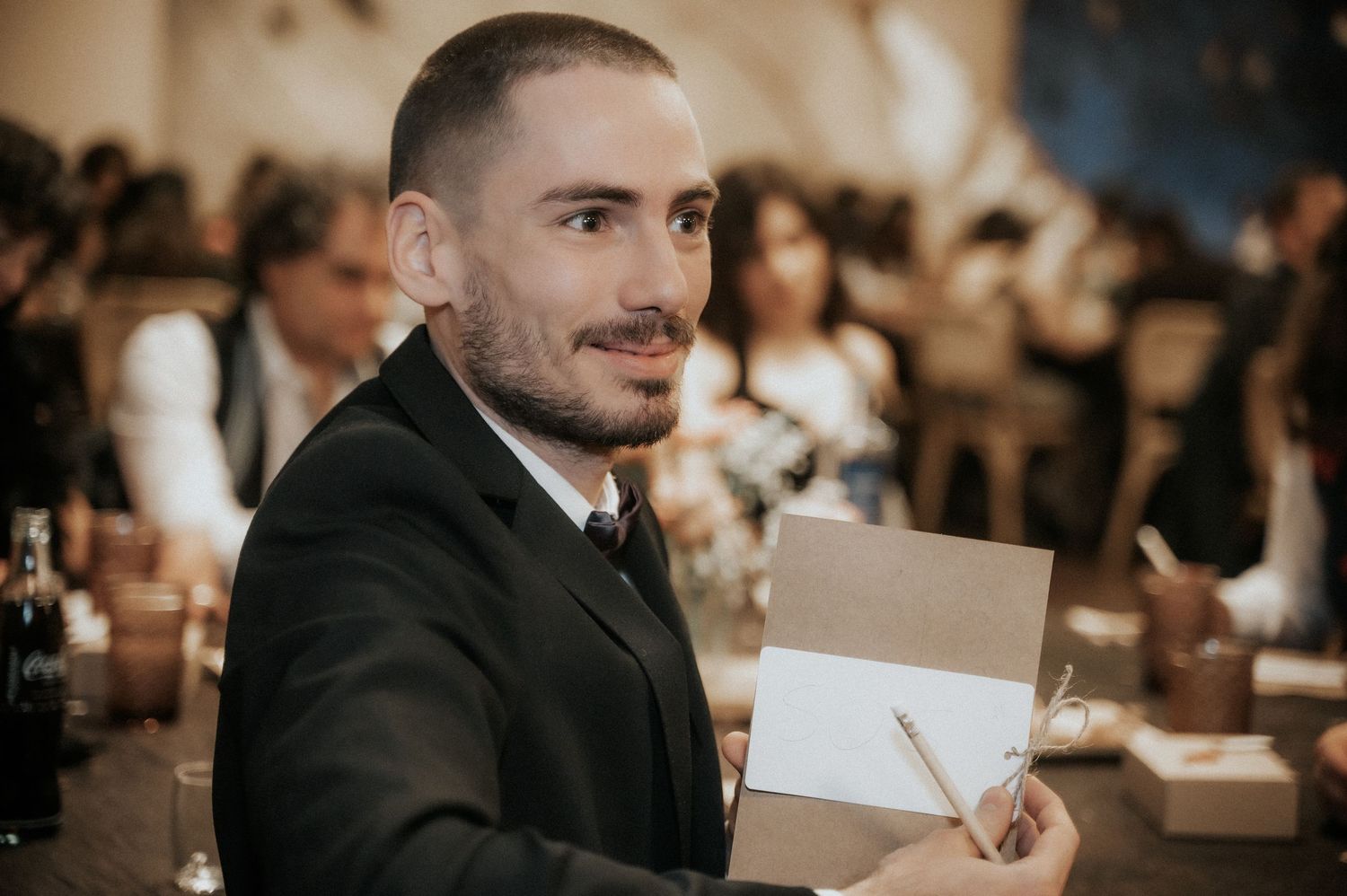 Wedding guests in formal attire smile and laugh while reading cards at a reception table.