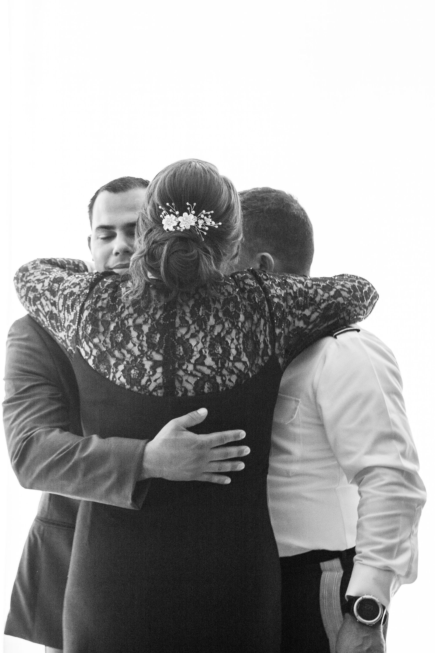 Black and white photo showing three people embracing in an emotional group hug, with delicate floral hair details visible.