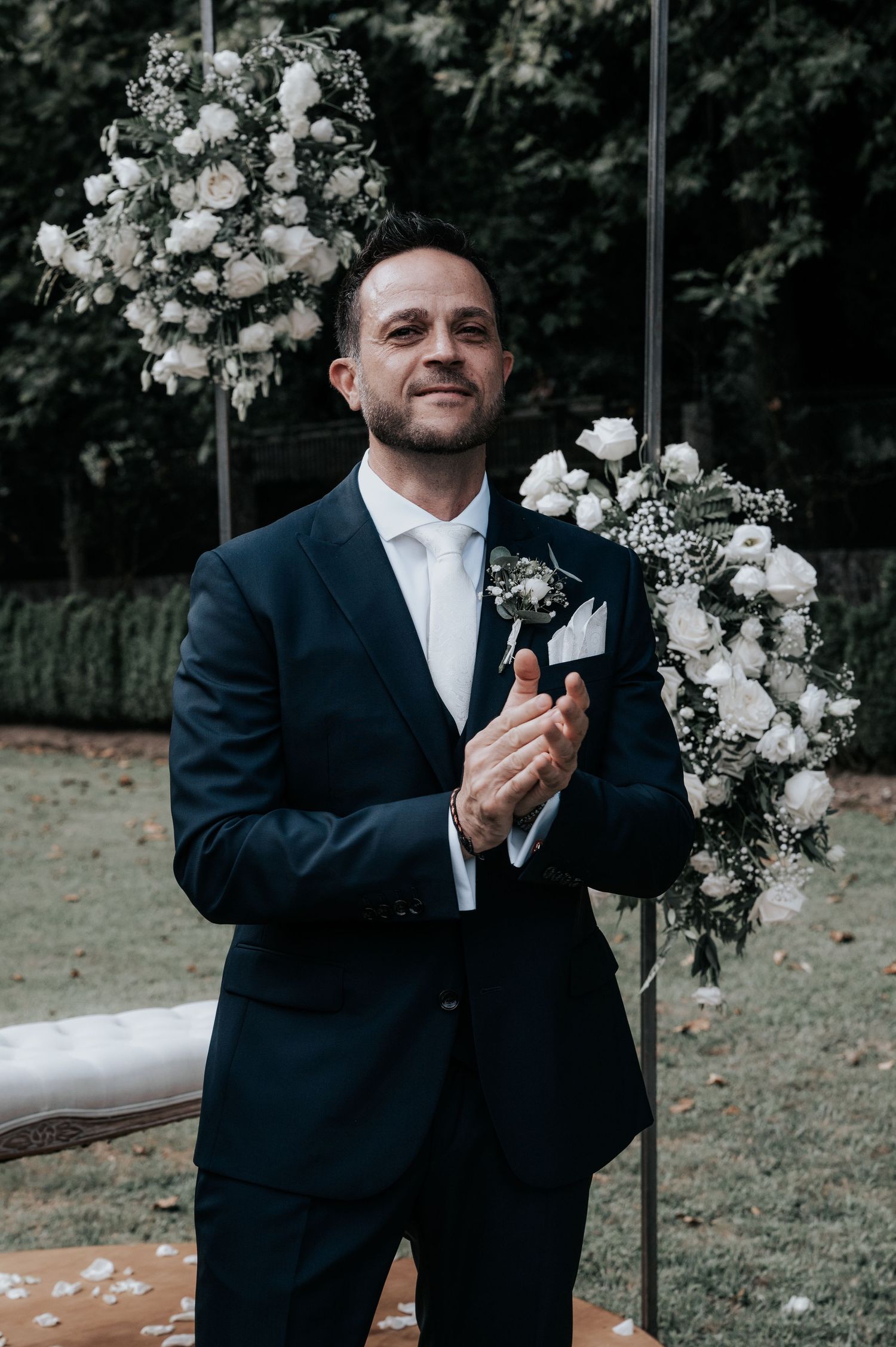 A groom in a navy suit and white tie adjusts his boutonniere next to elegant white floral arrangements outdoors.
