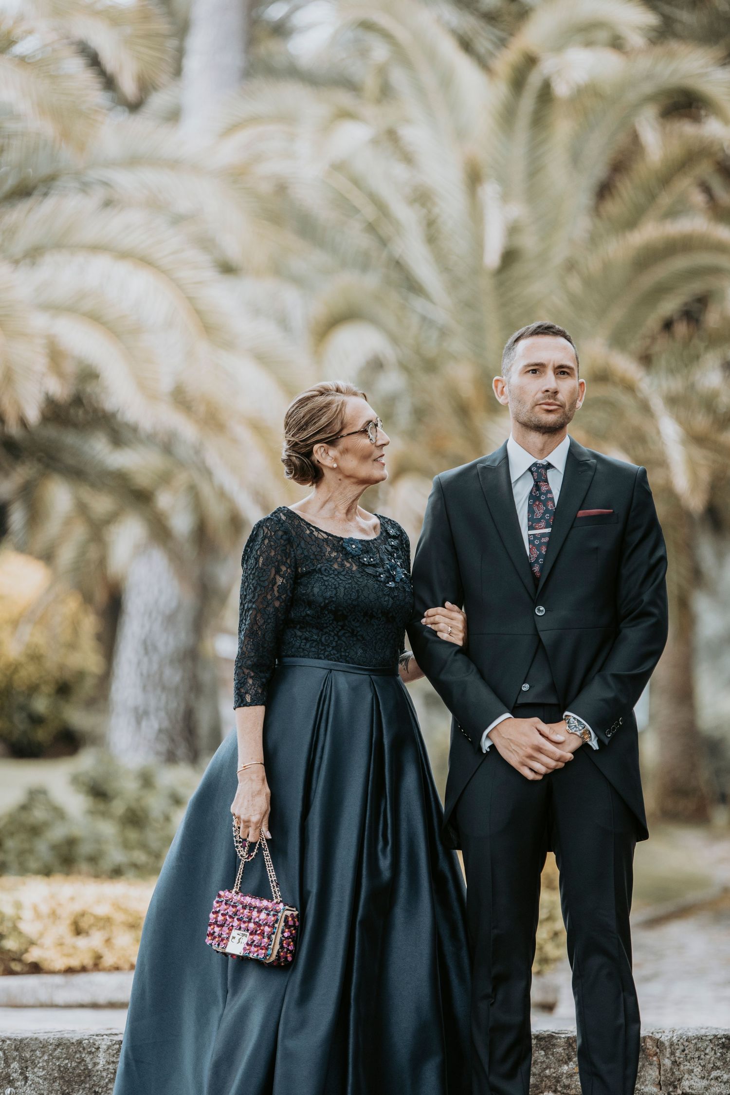 Couple in formal black attire poses together in an outdoor garden setting with palm trees in the background.