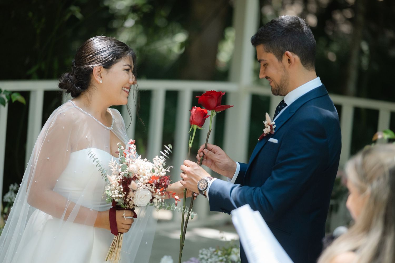 A couple in wedding attire shares a tender moment in a garden setting with roses and white flowers.