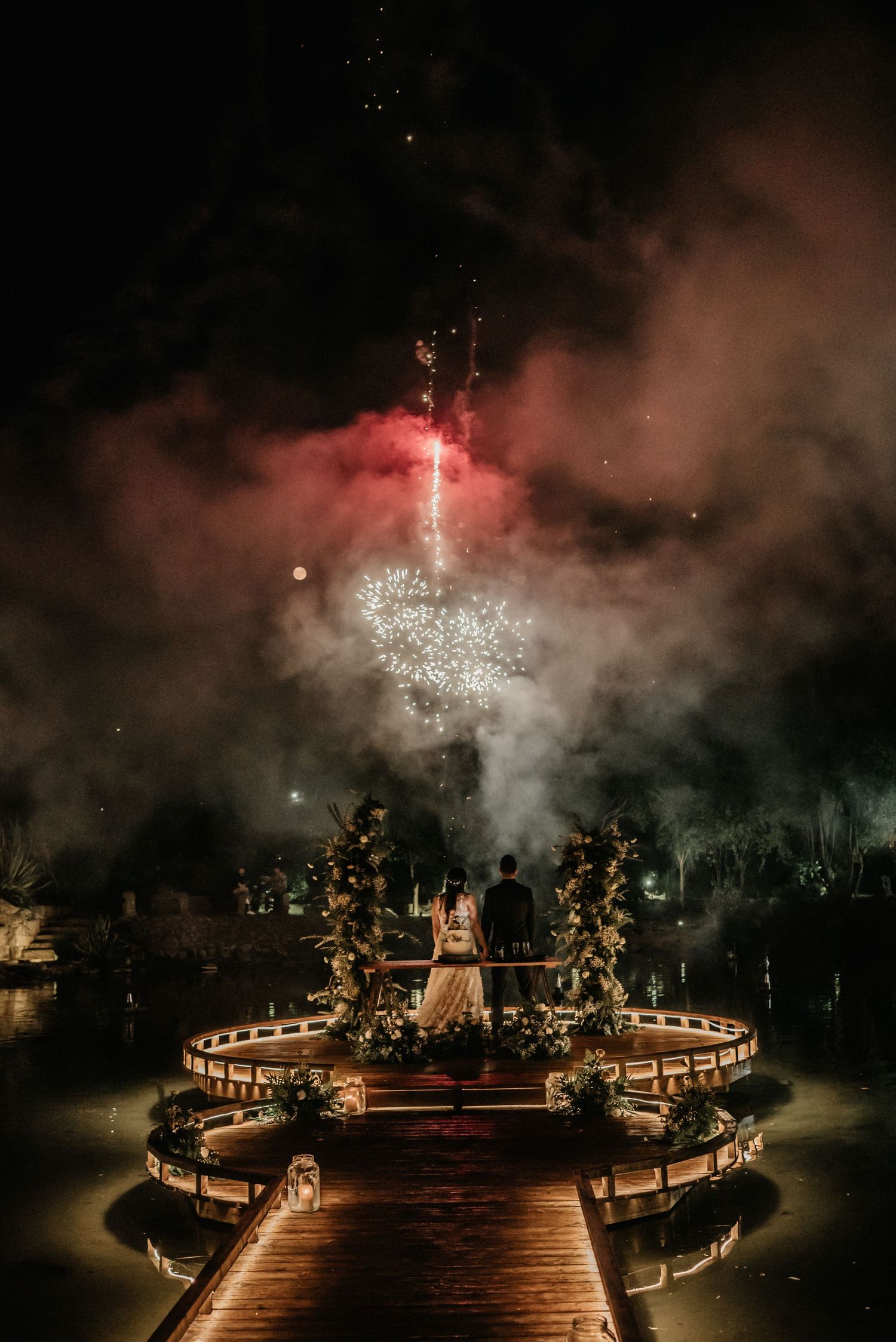 Fireworks illuminate a circular outdoor venue with glowing lights and smoke creating a dramatic nighttime atmosphere.