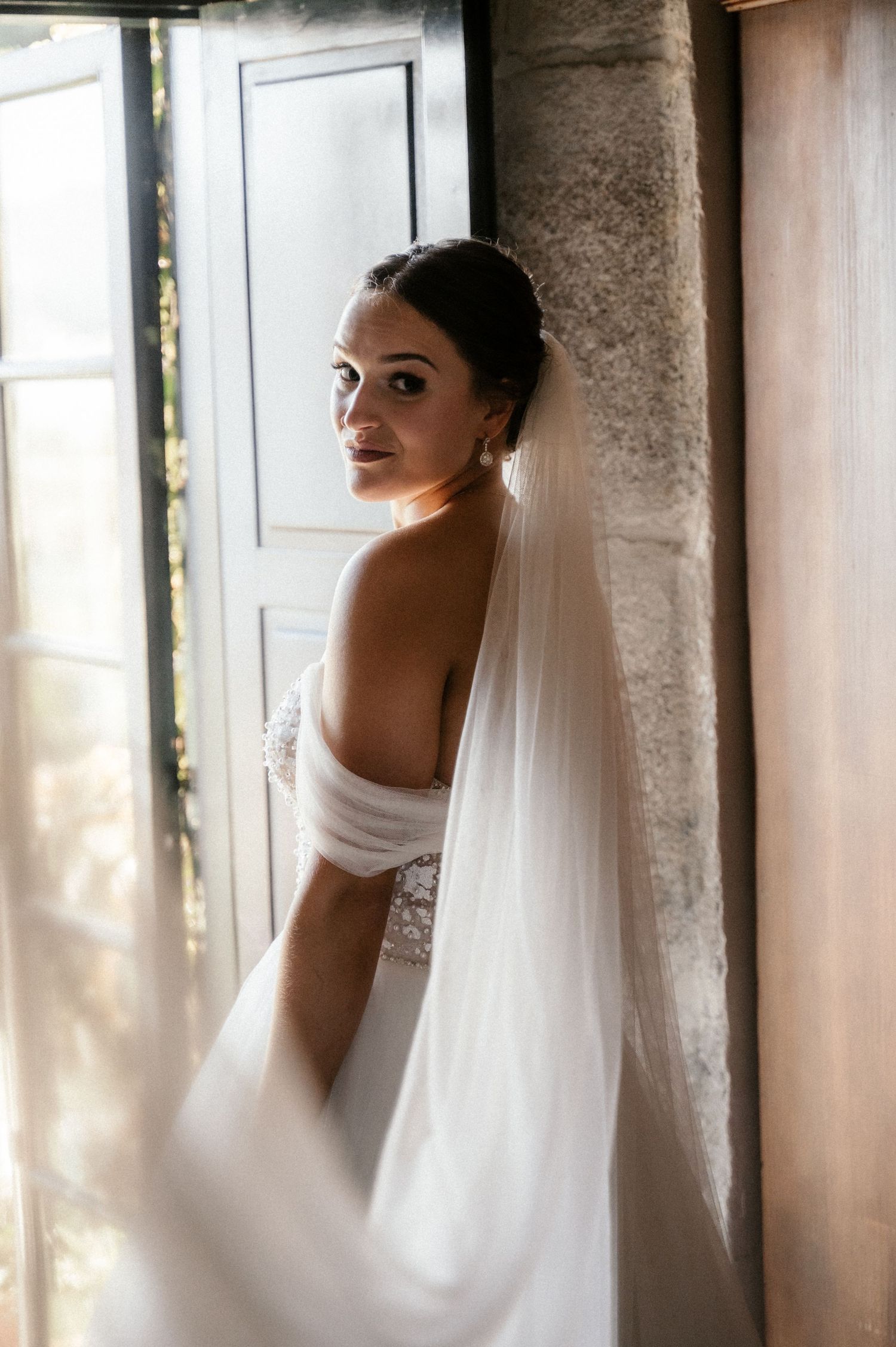 A bride in a classic white wedding dress and veil poses elegantly by a rustic doorway with natural lighting.
