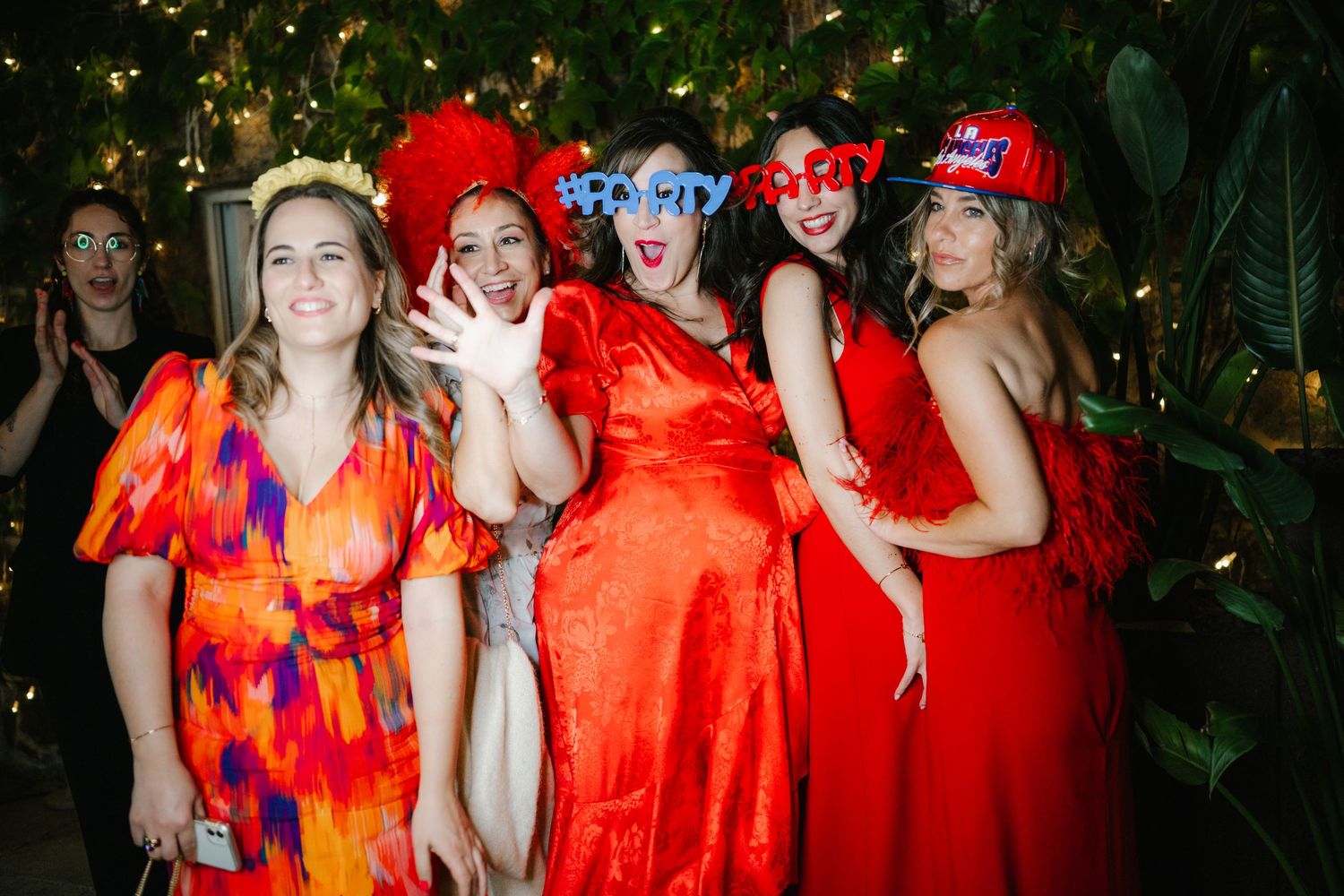 A group wearing vibrant red and orange dresses pose together during a festive celebration with twinkling lights.
