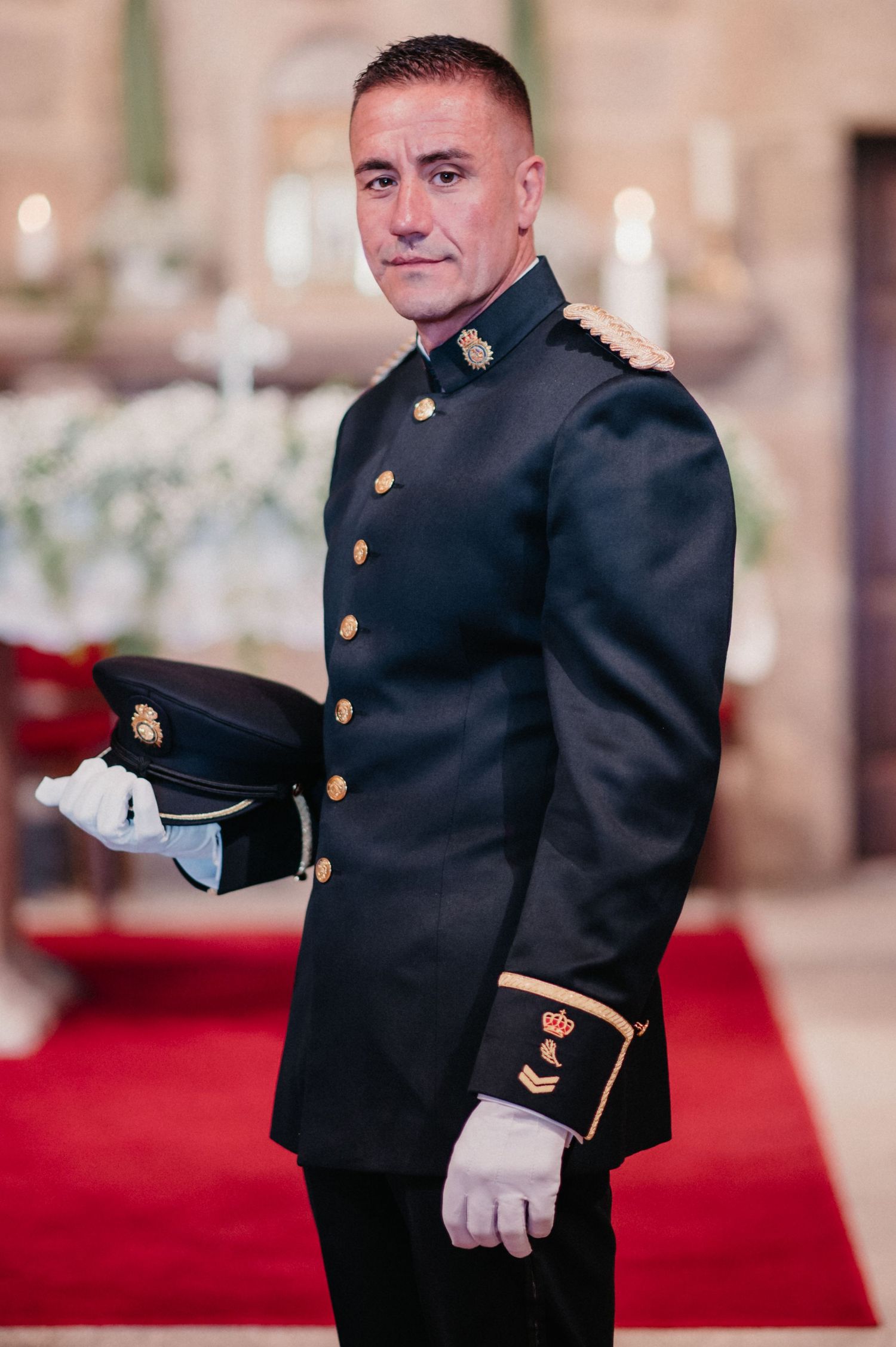 Military service member in formal dress uniform with brass buttons stands at attention on red carpet.