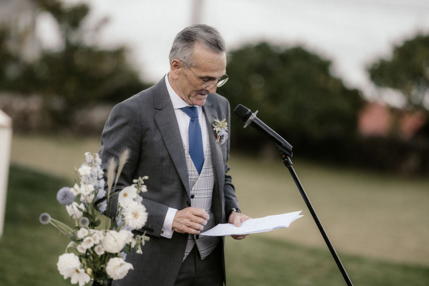 A person in a gray suit gives an emotional speech at an outdoor wedding ceremony.