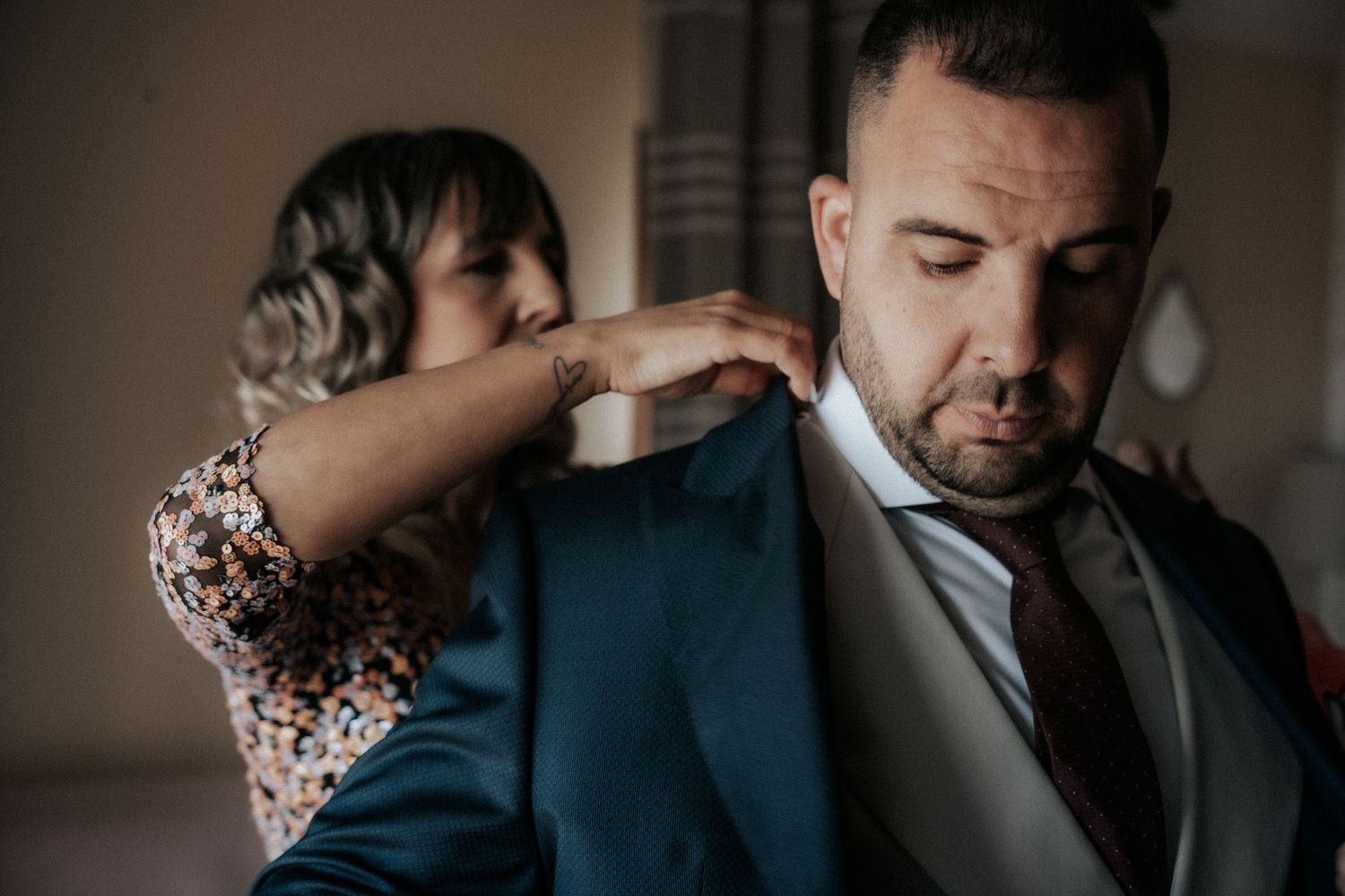 A person helps adjust the collar of someone wearing a navy suit and tie in a dimly lit room.