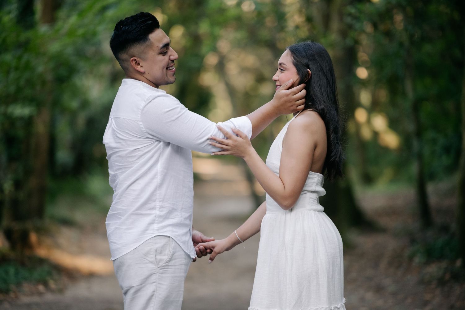 A romantic couple in white outfits share an intimate moment on a wooded path during a golden hour photoshoot.