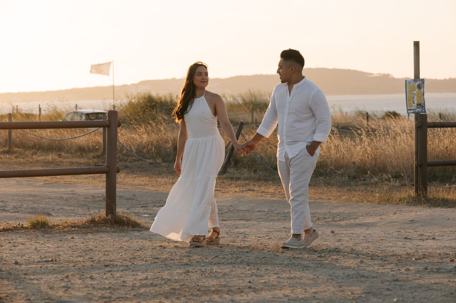 A couple in white outfits walks together through a scenic desert landscape at sunset.