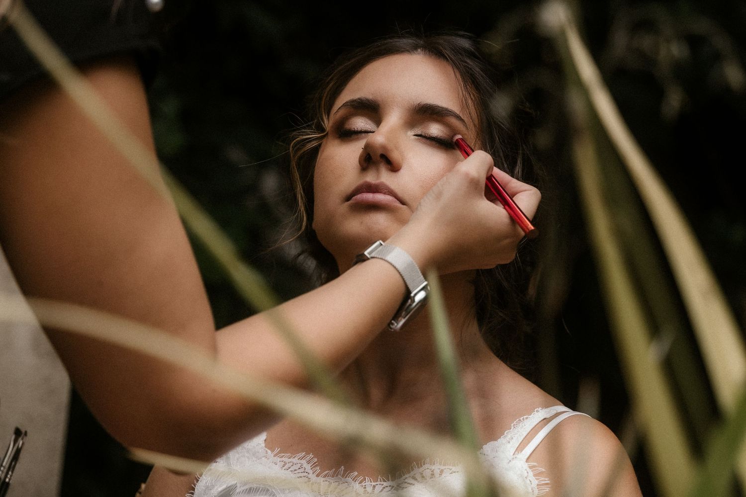 Makeup artist applying cosmetics on model in white dress with natural lighting and foliage background.