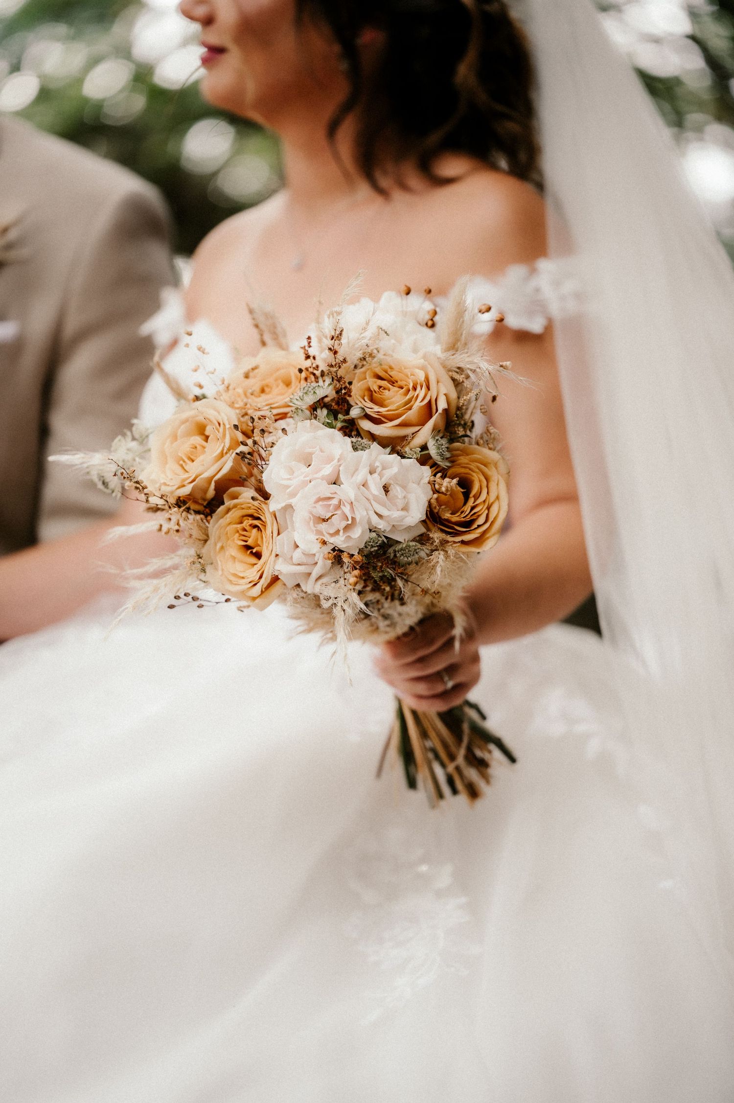 Close-up view of a romantic peach and blush wedding bouquet with roses and delicate dried elements against a white dress.