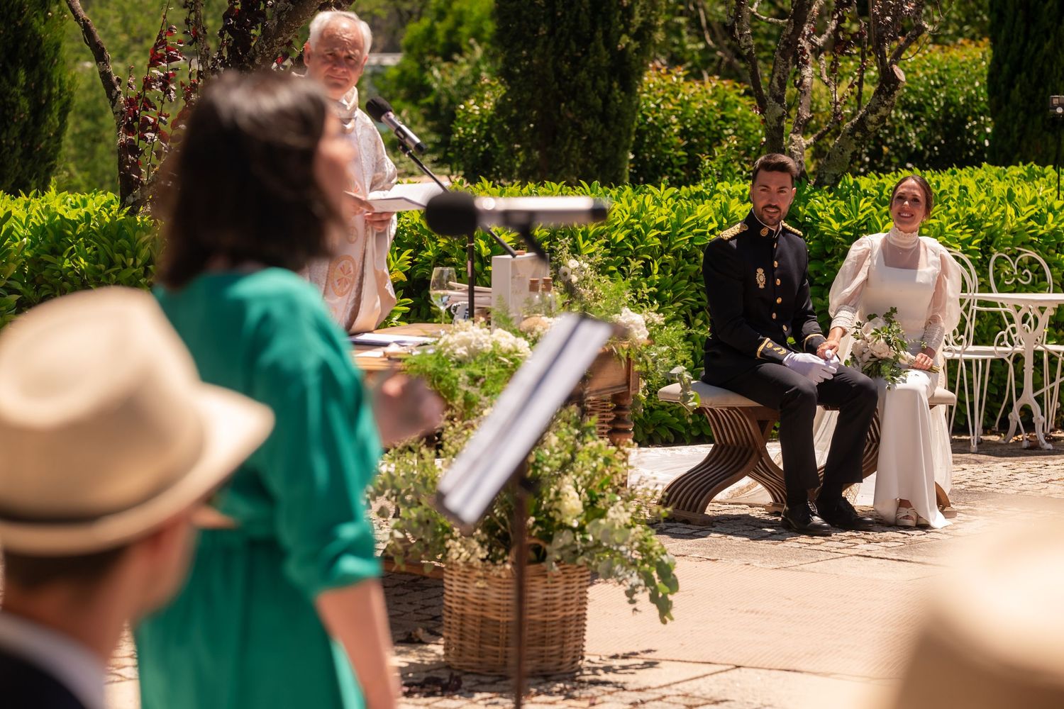 Wedding ceremony guests watch the proceedings from garden chairs in a sunny outdoor setting.