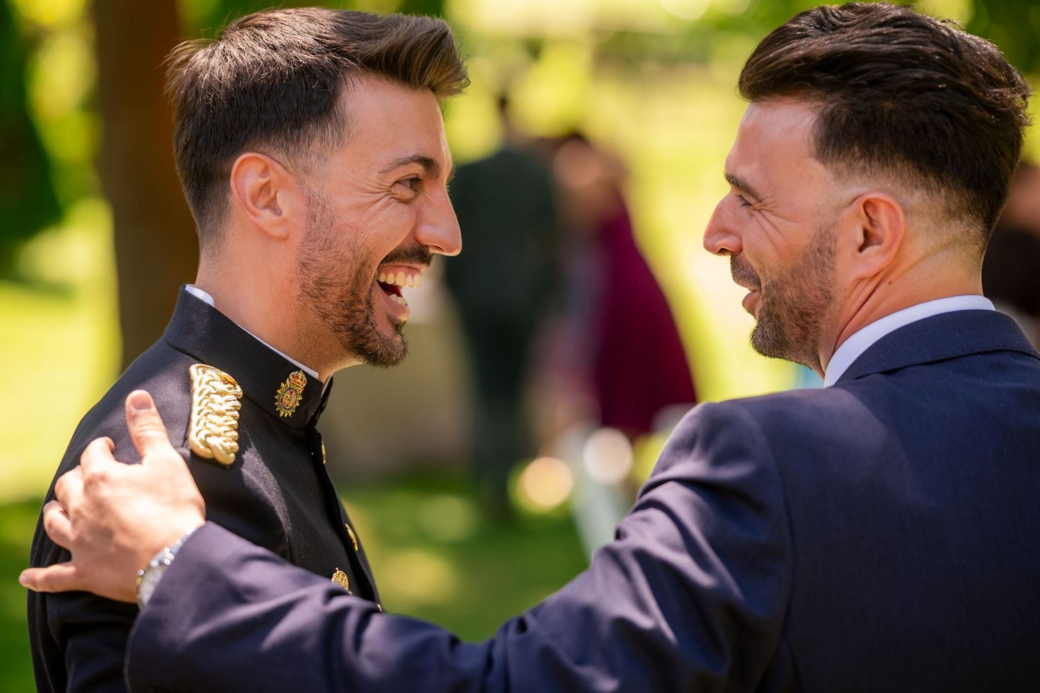 Two people in navy suits share a laugh during an outdoor event in bright sunlight.