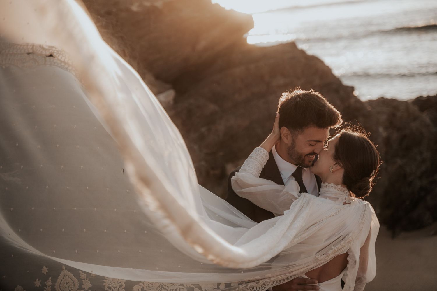 Ethereal wedding photo with flowing veil catching wind as newlyweds embrace during golden hour.