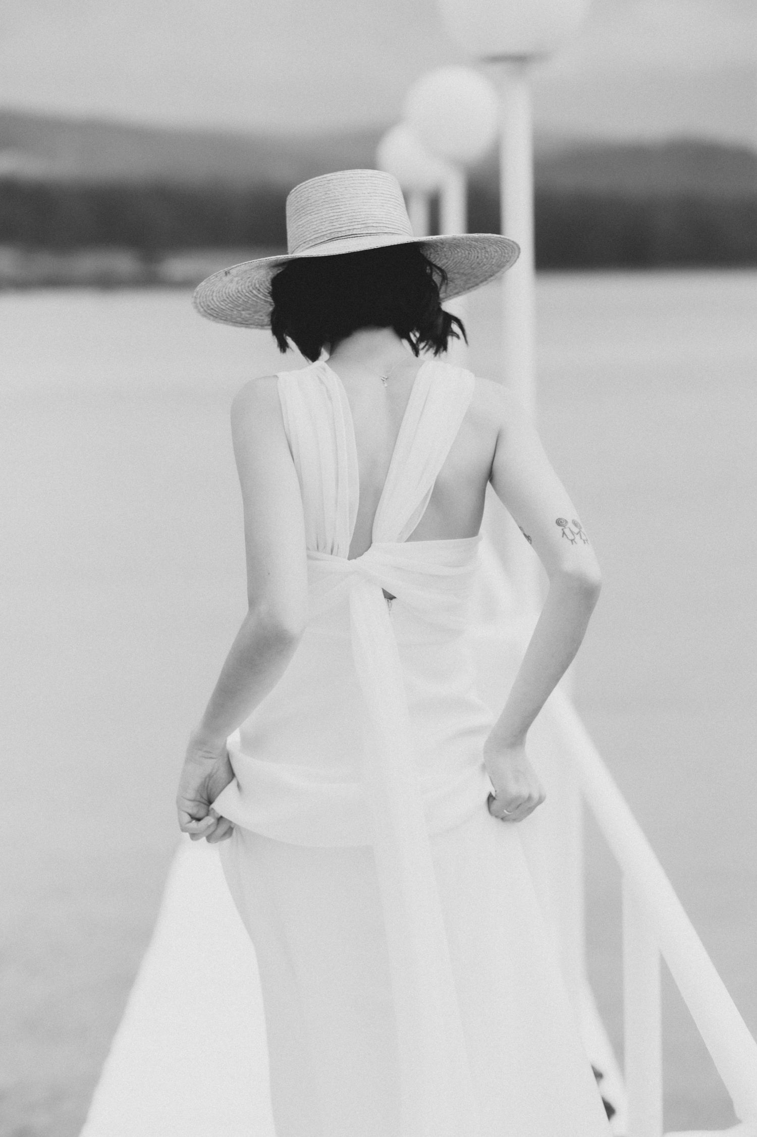 A figure in an ethereal white dress and sun hat stands gracefully by the water on a bright summer day.