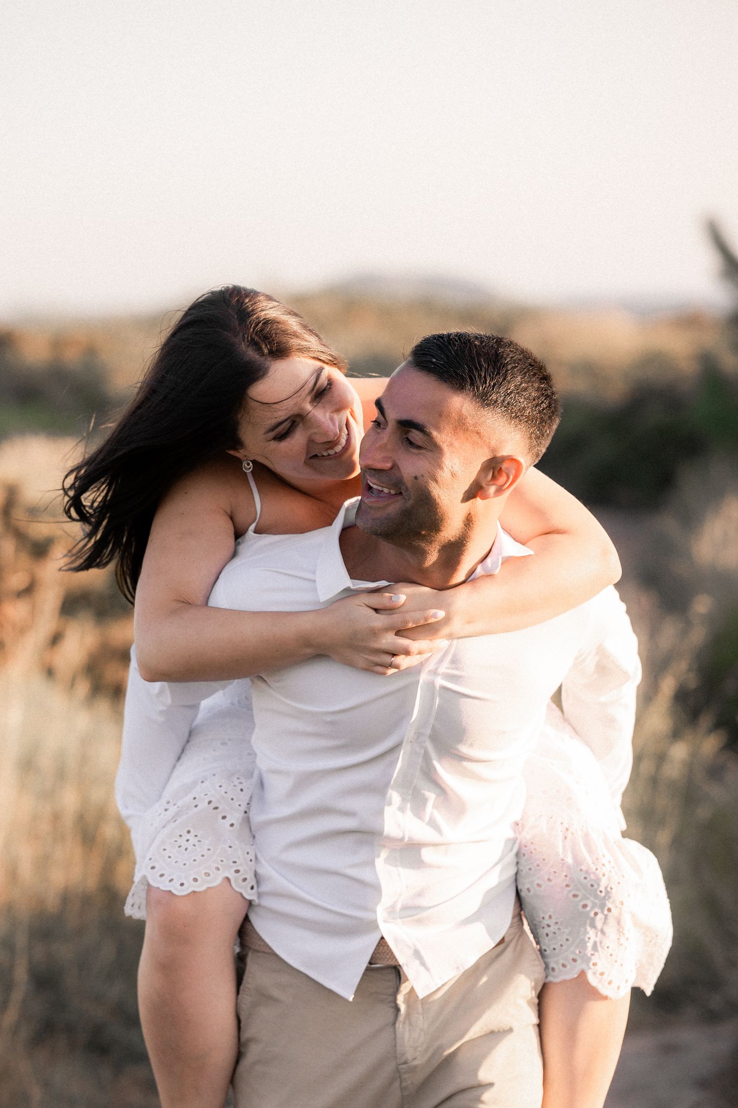 Romantic couple embracing in golden sunlight among tall grass during sunset photoshoot.
