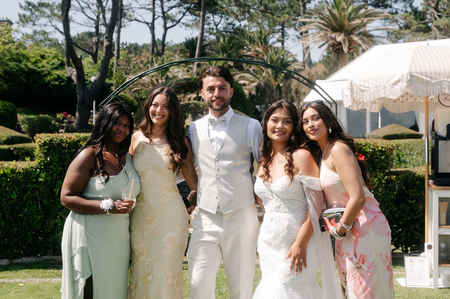 Wedding party in white and pink attire poses together outdoors with palm trees and greenery in the background.