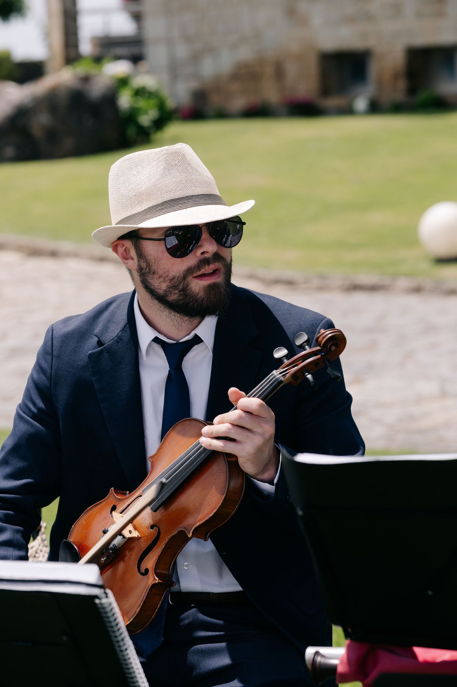 A musician in a dark suit and hat plays violin outdoors during a daytime performance.