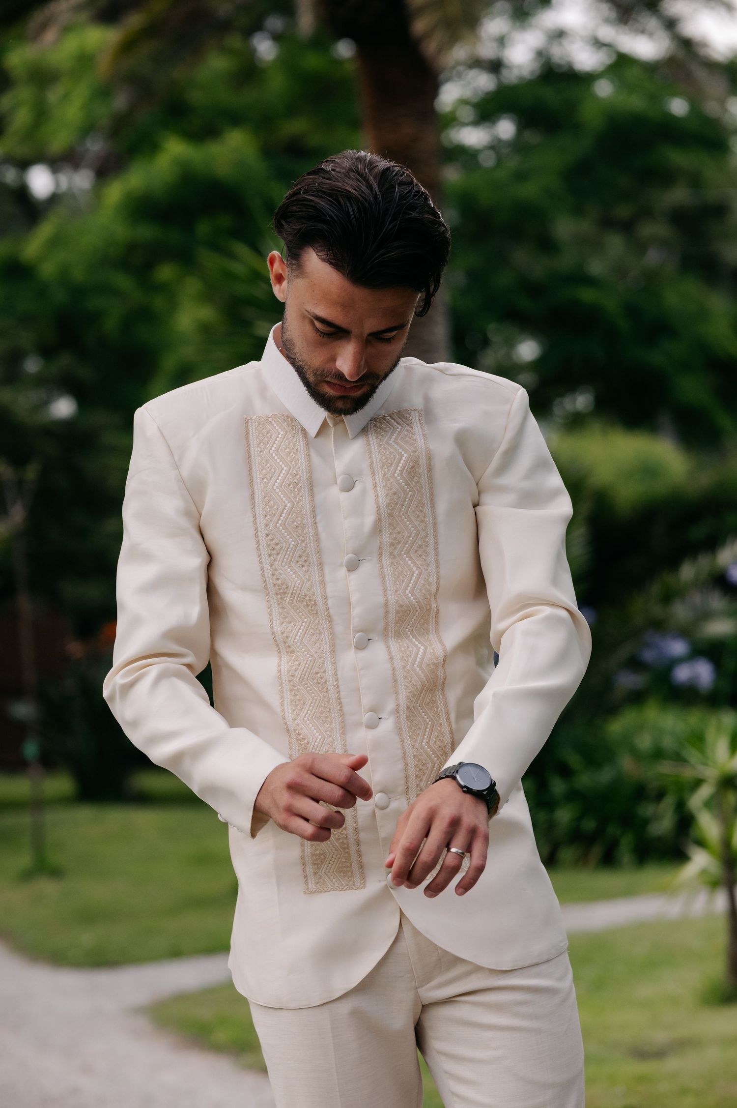 A person in a cream-colored traditional barong tagalog formal suit checks their watch while standing in a garden setting.