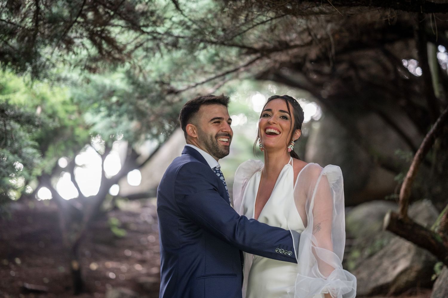 Romantic wedding couple embraces in a sunlit garden setting with natural greenery and trees in the background.
