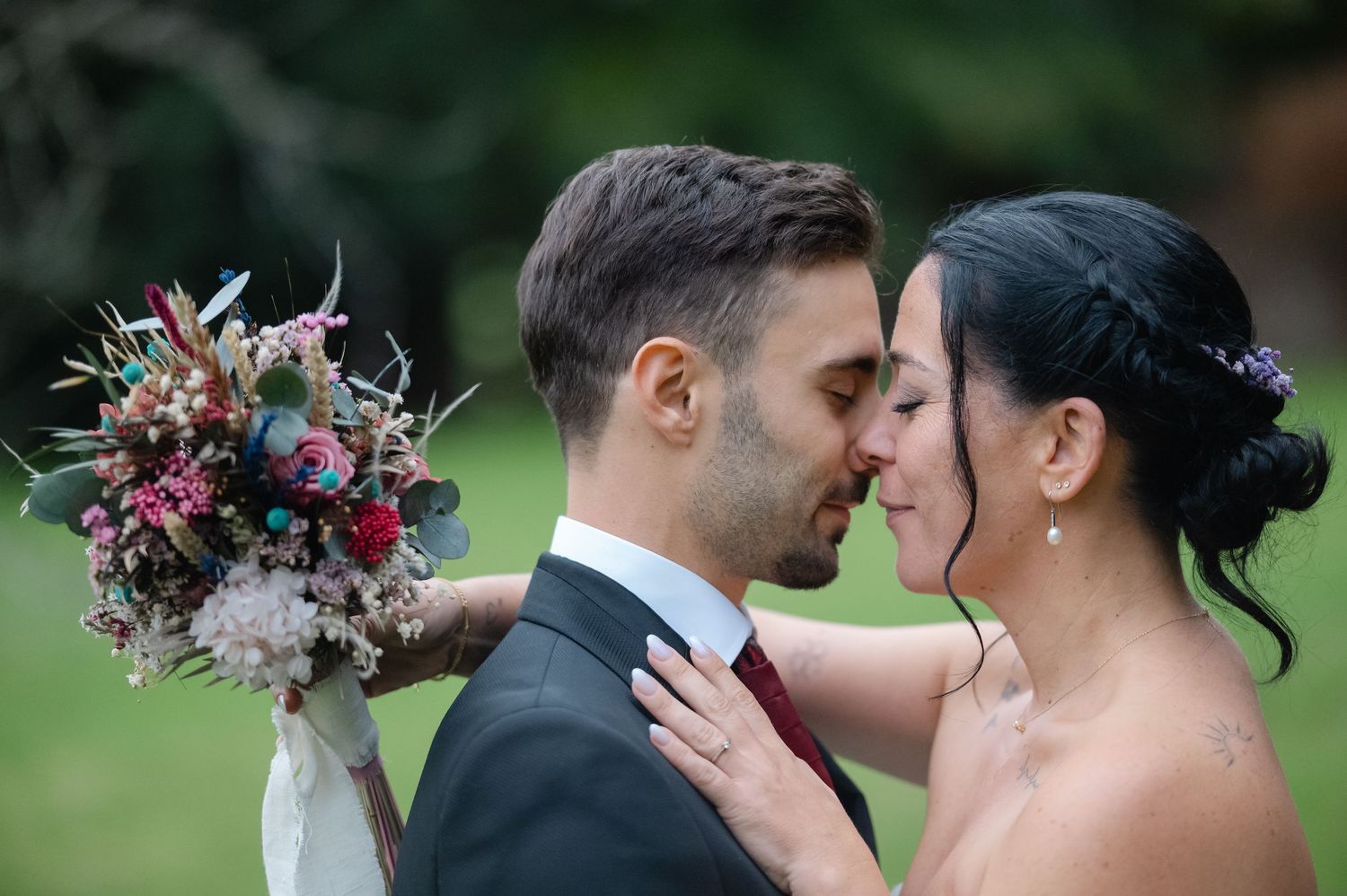 A romantic wedding couple embraces while holding a vibrant burgundy and pink floral bouquet in an outdoor garden setting.