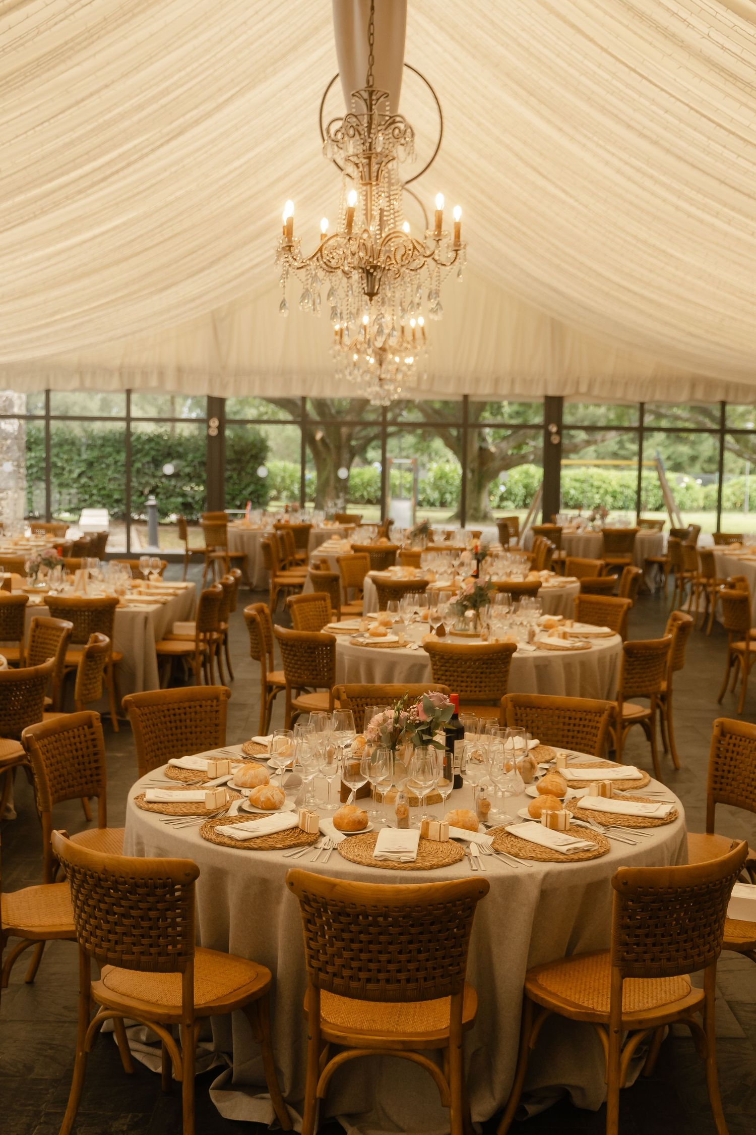 Elegant wedding reception setup under white tent with chandeliers and round tables set with fine dinnerware.