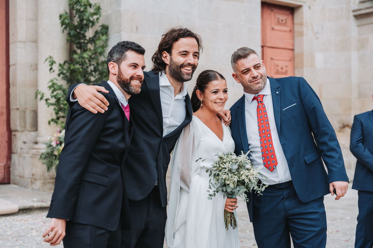 Wedding party members share joyful laughter together during outdoor photo session in front of stone building.