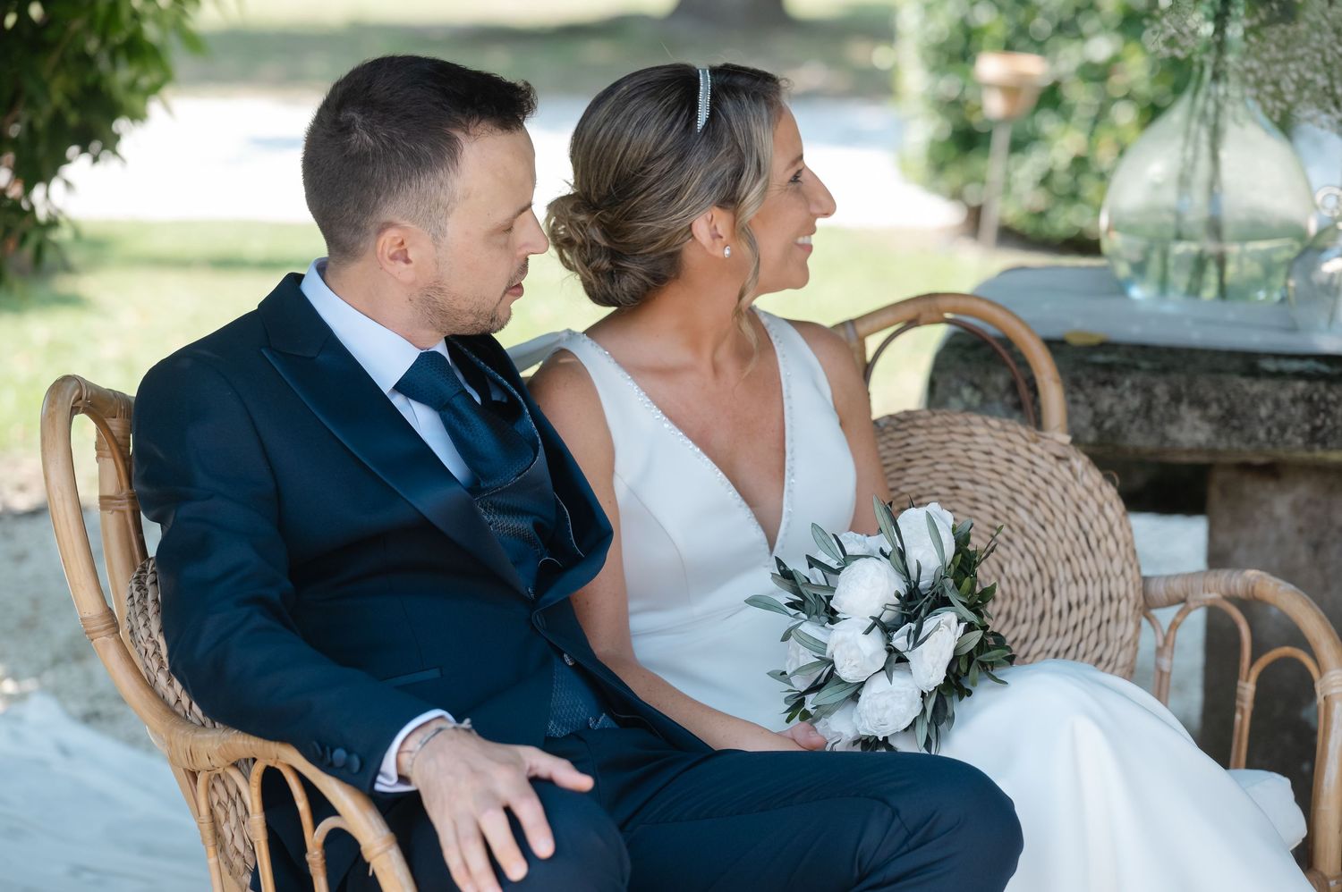 A couple sits together on an elegant vintage chair during an outdoor wedding celebration.