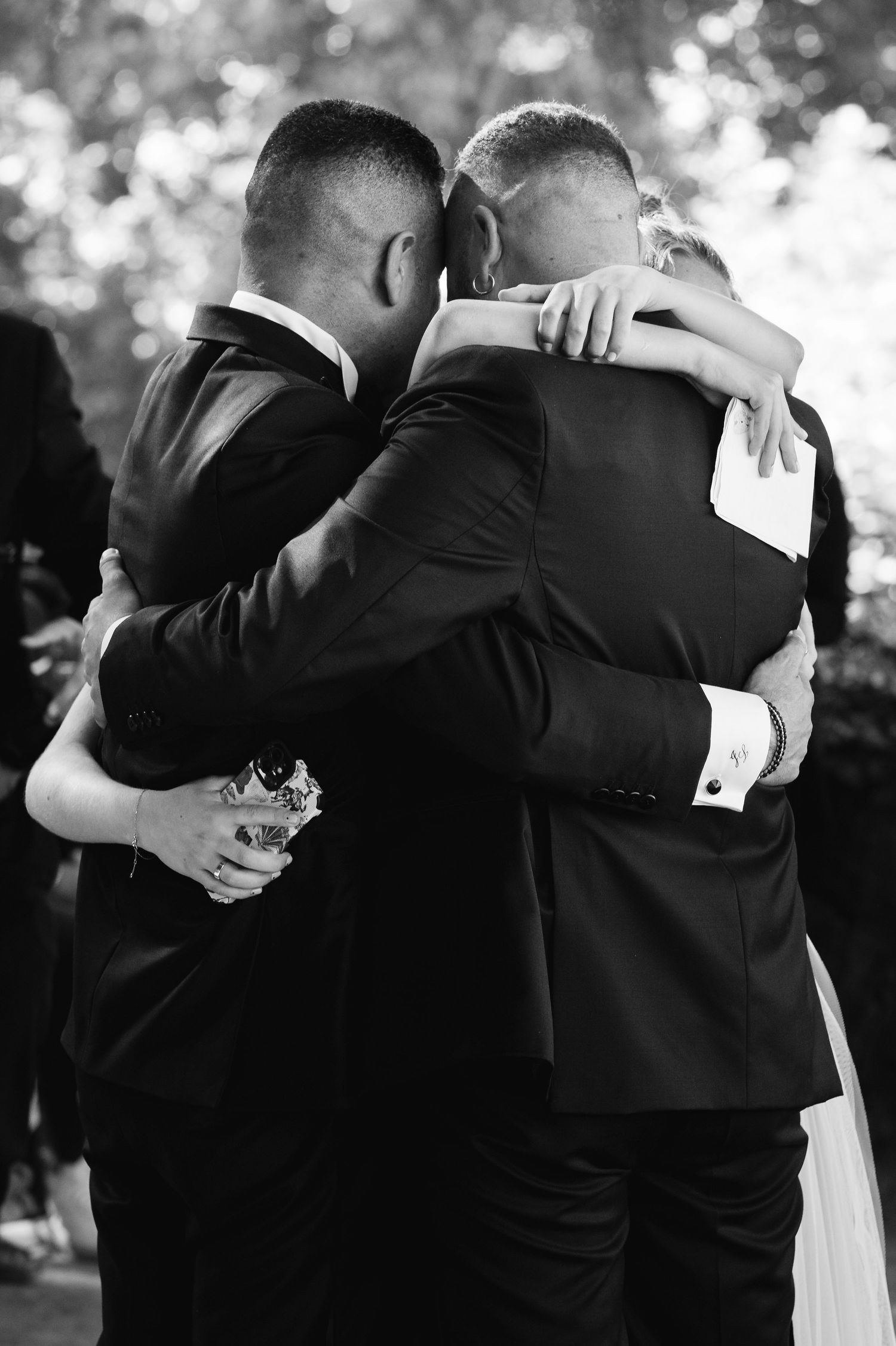 Black and white photo of three people in suits sharing an emotional embrace during a wedding ceremony.