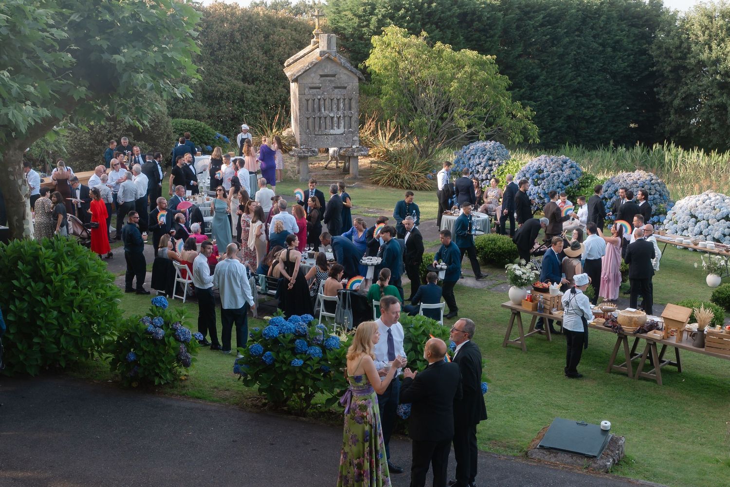 A large outdoor wedding reception gathering in a lush garden setting with guests mingling during golden hour.