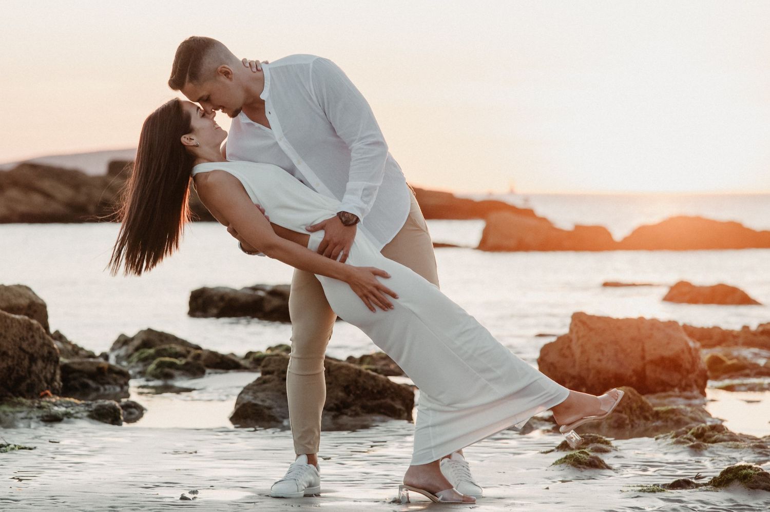 Romantic couple embracing on rocky beach during golden sunset with waves crashing around them.