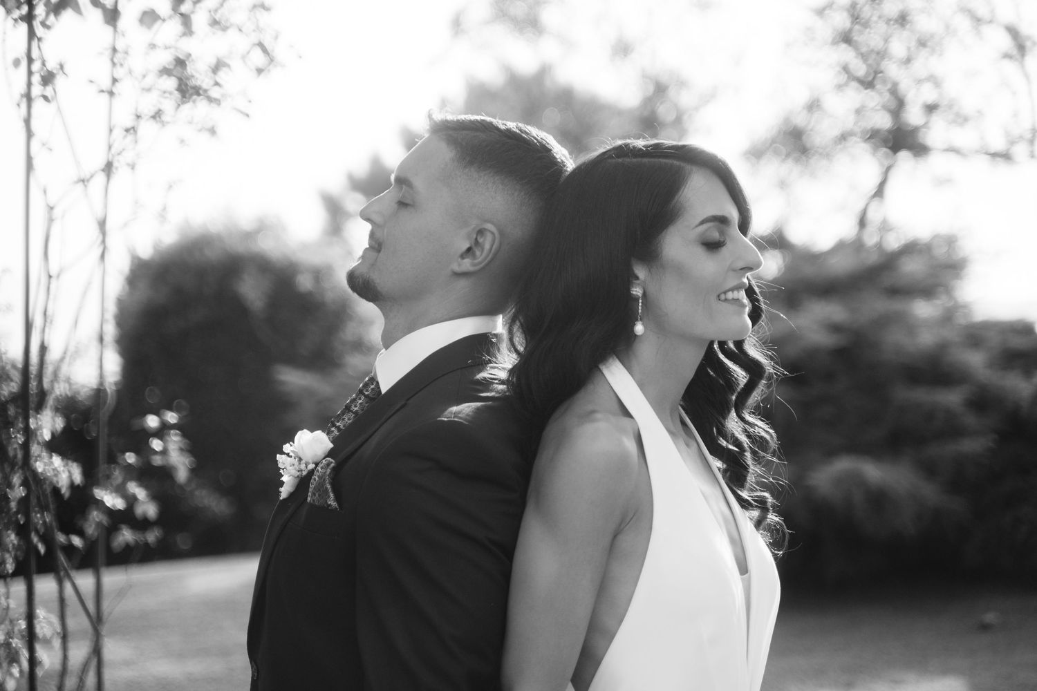 A couple in wedding attire stand back to back outdoors in a black and white photograph.