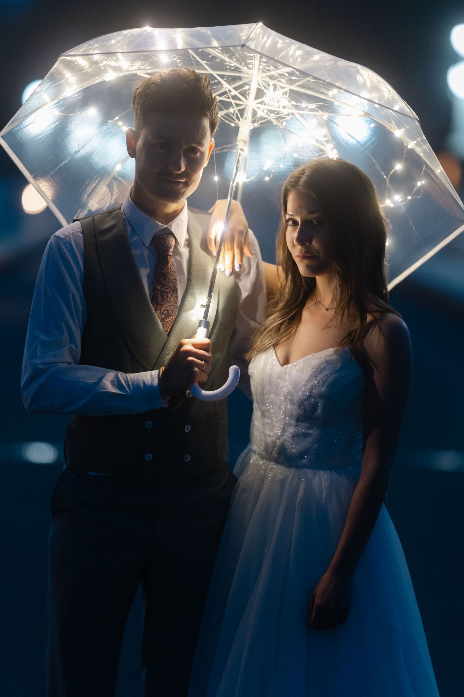 Bride and groom standing under a transparent umbrella with fairy lights at night, creating a romantic wedding portrait.