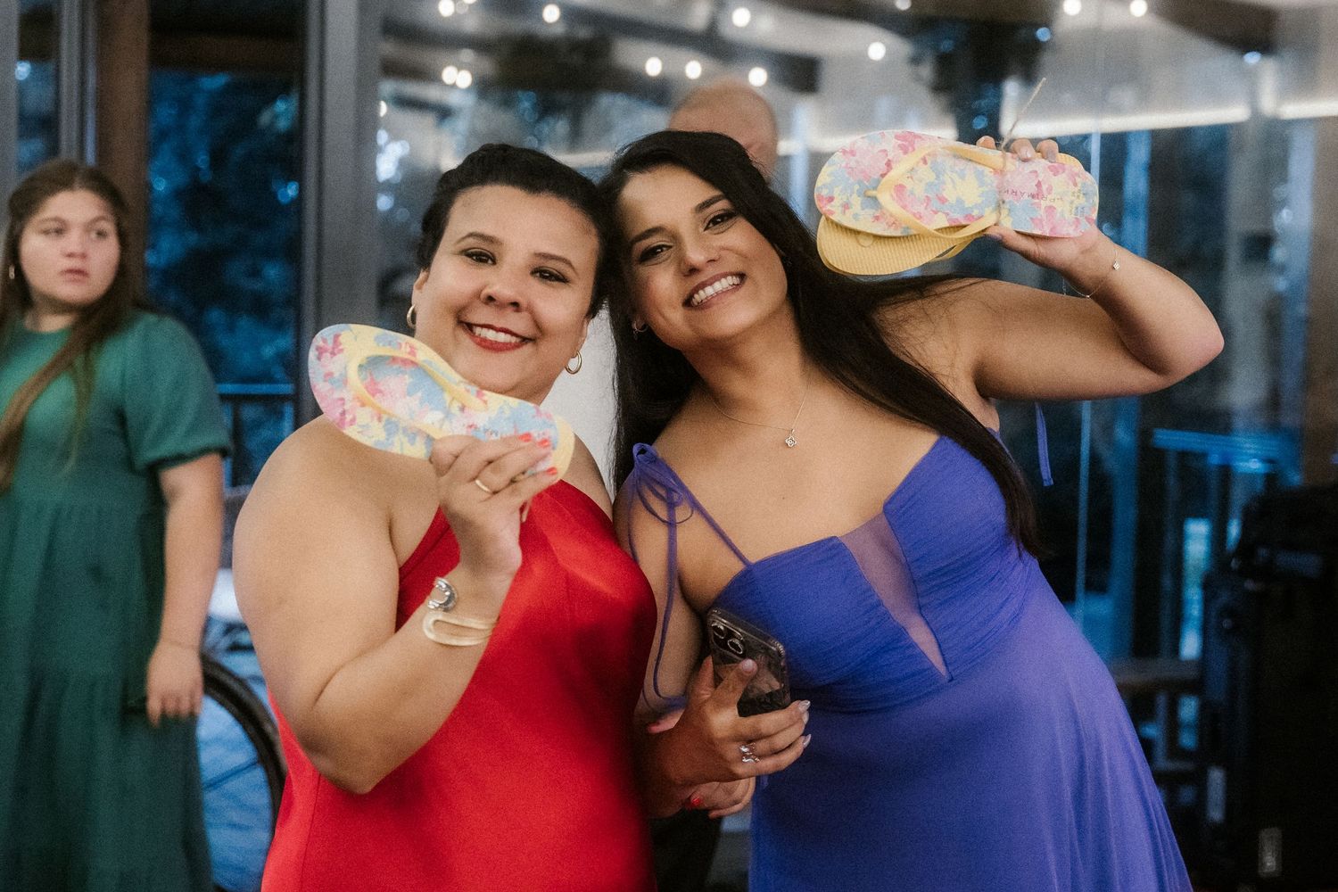 Two friends in red and blue dresses hold up matching decorated cakes at an evening celebration.