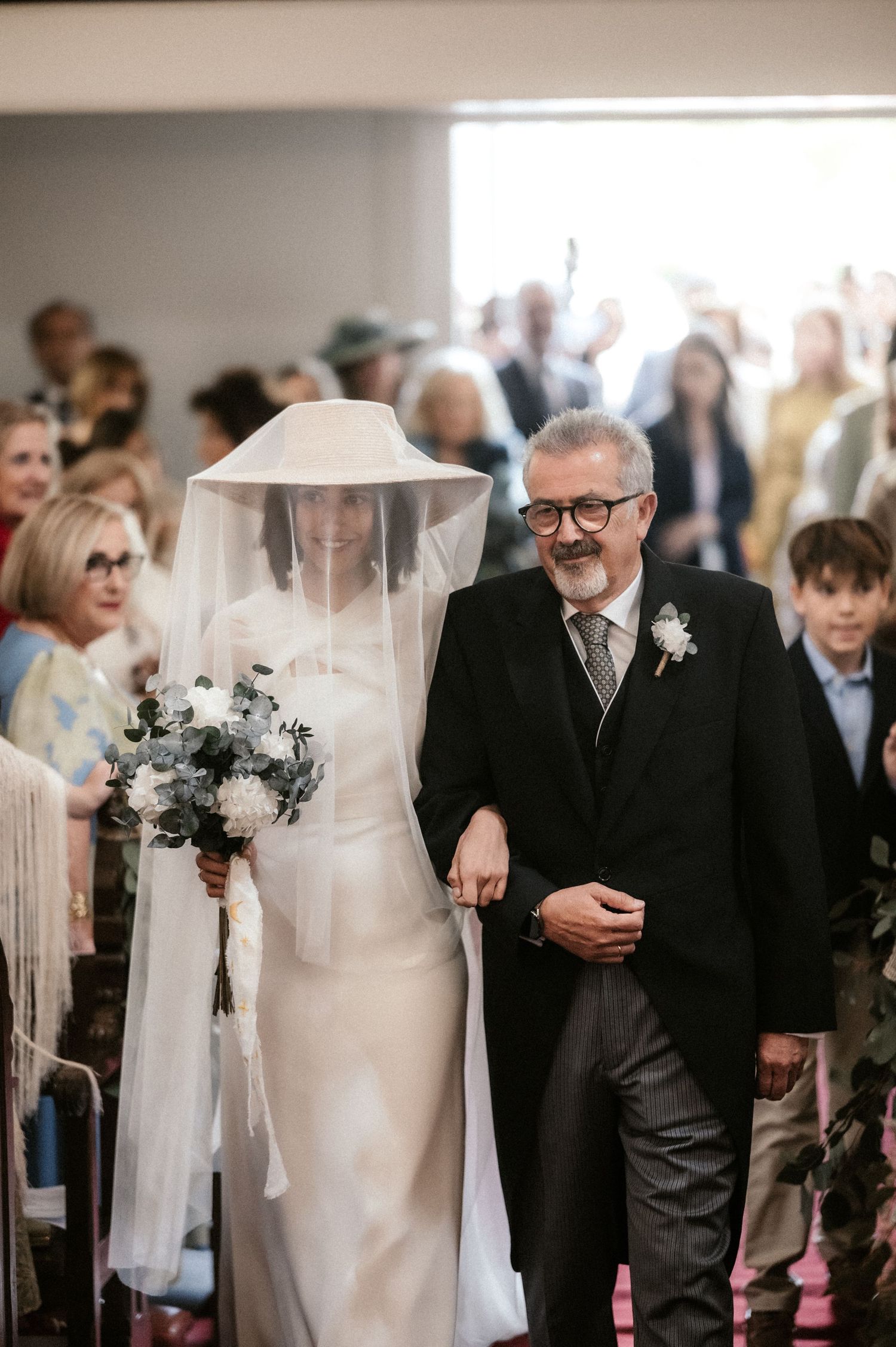 A bride in an elegant white dress and wide-brimmed hat veil walks down the church aisle with her escort.