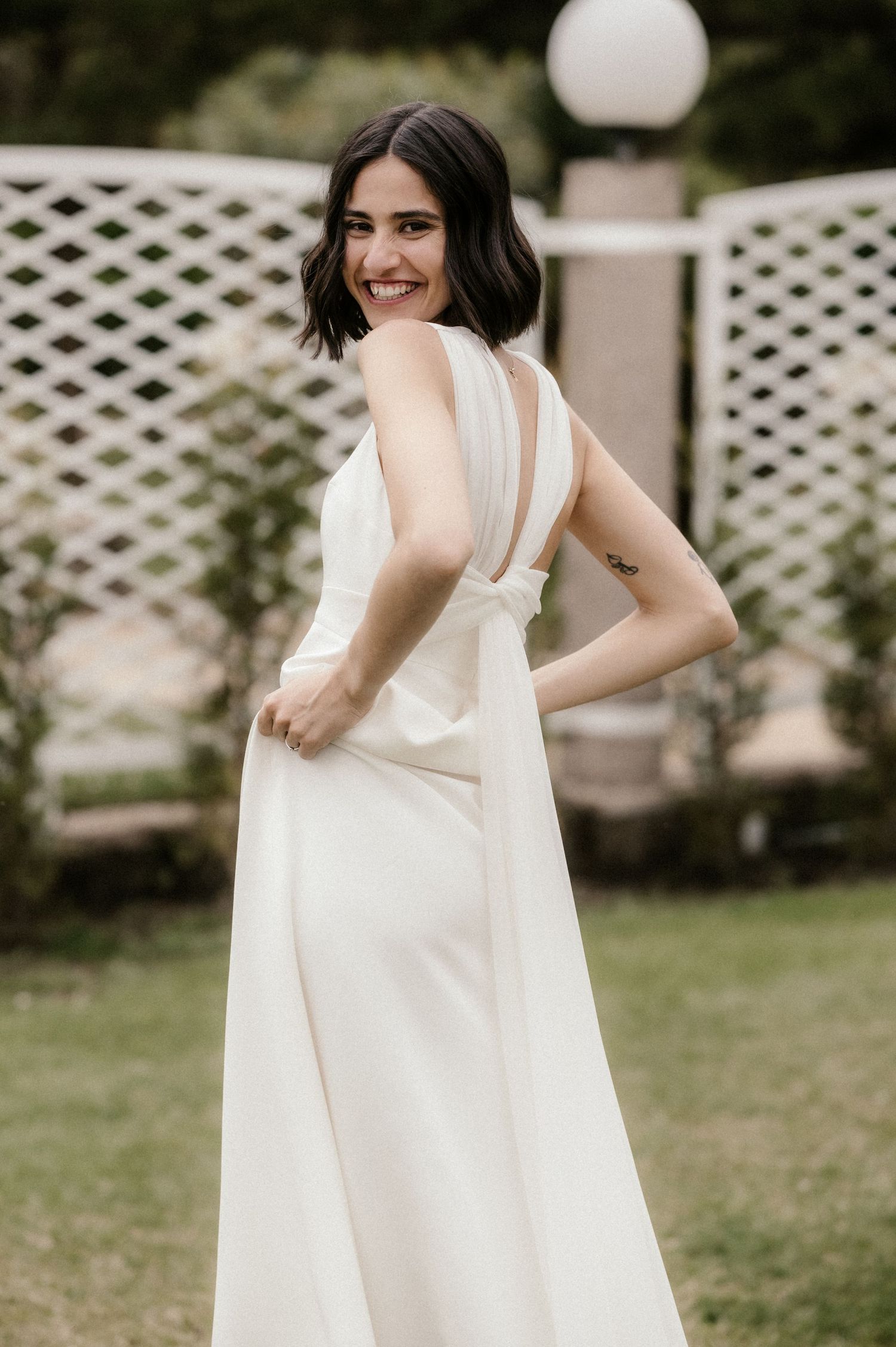 An elegant white wedding dress with unique back detail and straps shown in an outdoor garden setting.