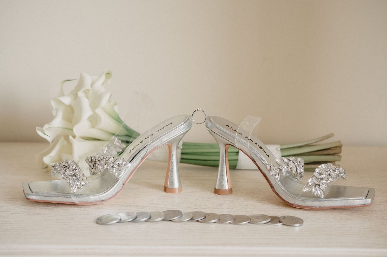 Elegant silver bridal shoes with crystal embellishments and high heels displayed next to white flowers.