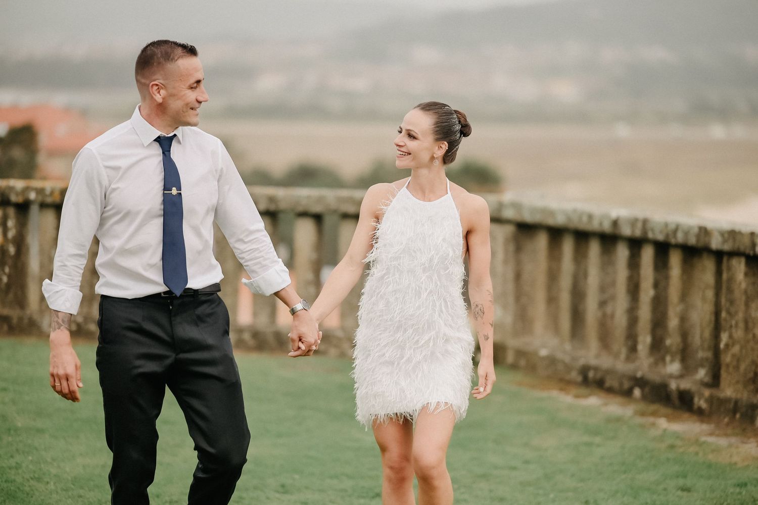A couple walks hand in hand along a scenic overlook with rolling hills in the background during a wedding celebration.