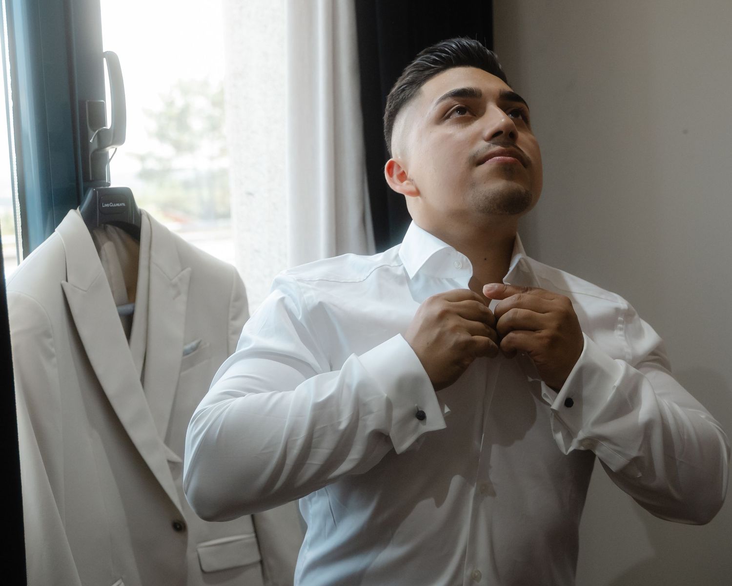 A person in a white dress shirt buttons up their collar while standing next to a light colored suit jacket.