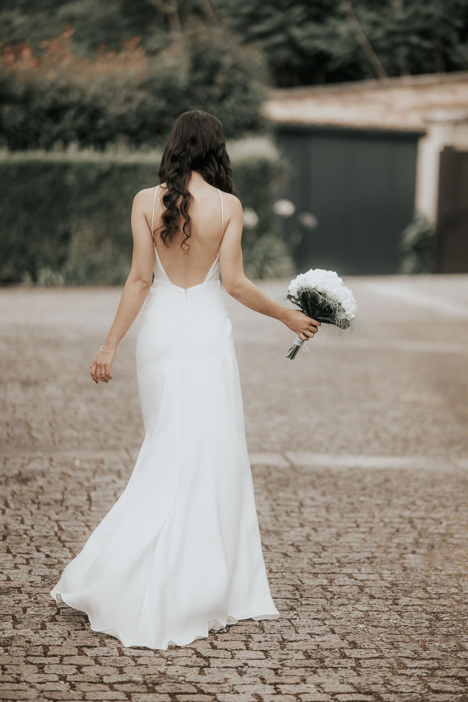 Back view of bride in elegant white backless wedding dress holding bouquet while walking on stone path outdoors.