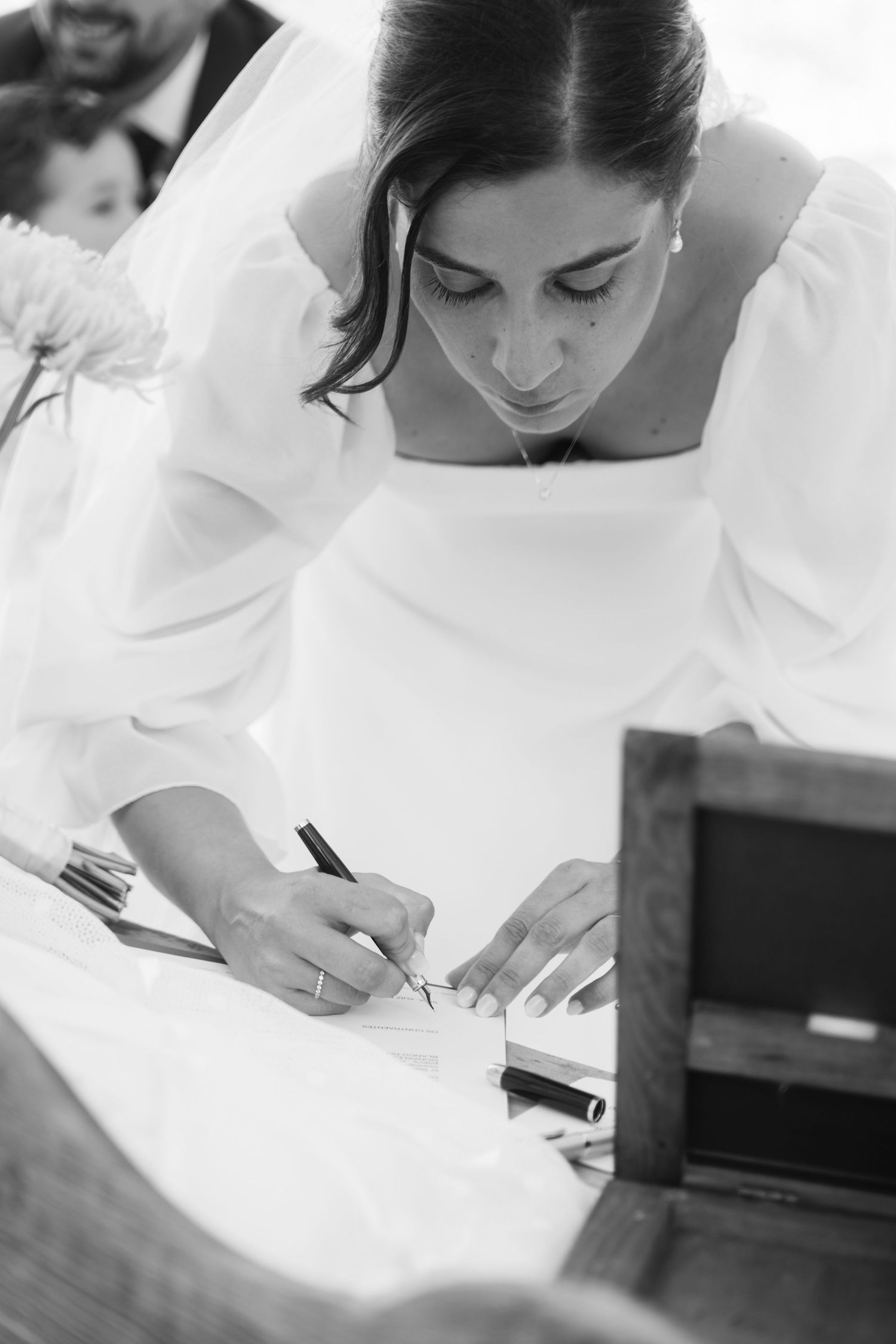 A bride in a white wedding dress leans over to sign documents during a wedding ceremony.