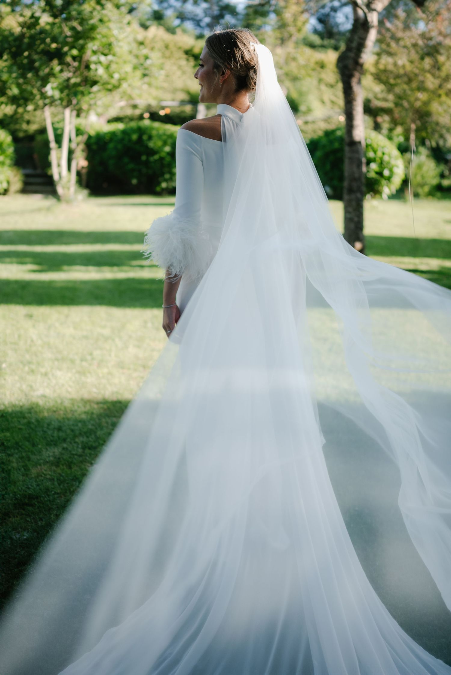 A bride in a flowing white wedding dress and veil stands in a sunlit garden setting.