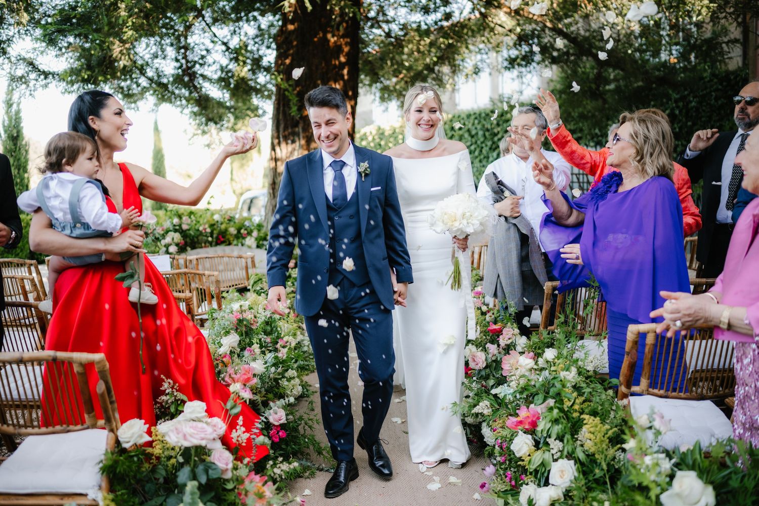 Wedding guests shower newlyweds with confetti as they walk down garden aisle surrounded by flowers.