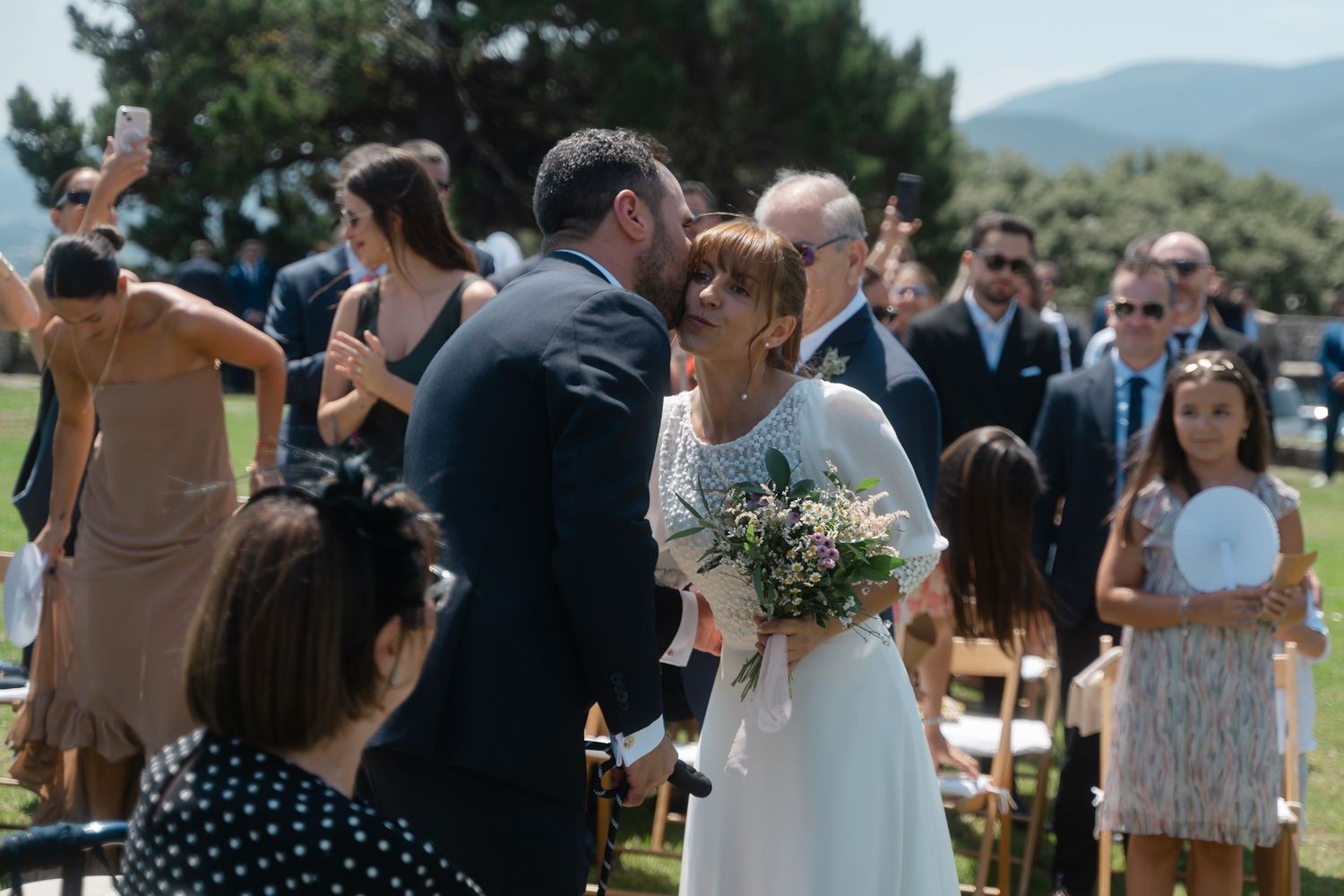 Outdoor wedding ceremony guests gather on a grassy hillside with mountains in the background.